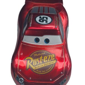 Peut inclure: Une voiture jouet rouge avec le num&eacute;ro 95 sur le toit et les c&ocirc;t&eacute;s. La voiture a des yeux bleus et un pare-brise blanc. L'avant de la voiture porte l'inscription "Rust-eze Bumper Ointment".