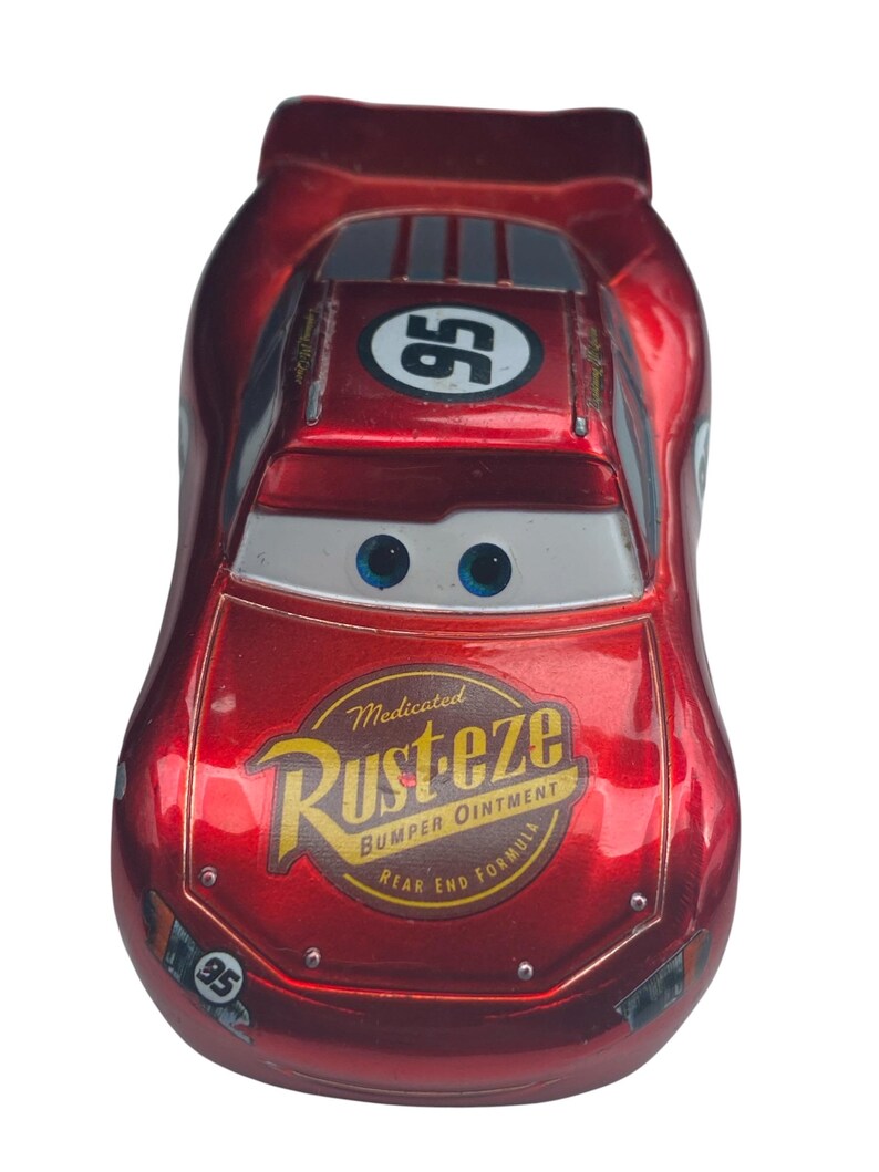 Peut inclure: Une voiture jouet rouge avec le num&eacute;ro 95 sur le toit et les c&ocirc;t&eacute;s. La voiture a des yeux bleus et un pare-brise blanc. L'avant de la voiture porte l'inscription "Rust-eze Bumper Ointment".