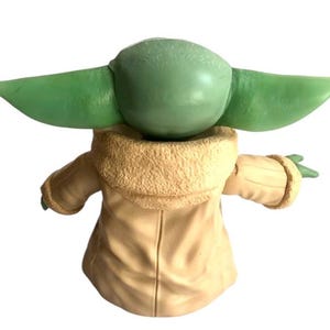 May include: A green and beige plush toy of a character from the Star Wars franchise. The toy is wearing a brown coat with a furry collar.