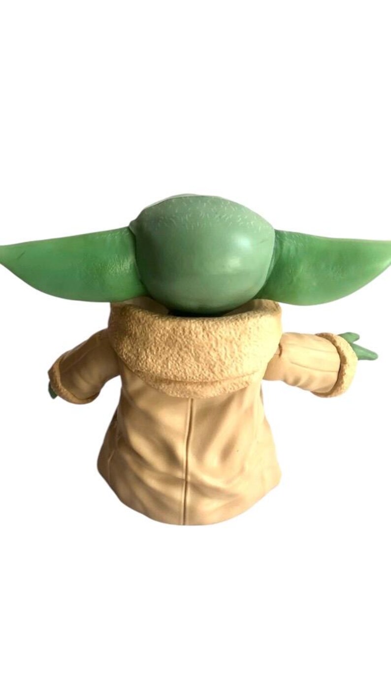 May include: A green and beige plush toy of a character from the Star Wars franchise. The toy is wearing a brown coat with a furry collar.