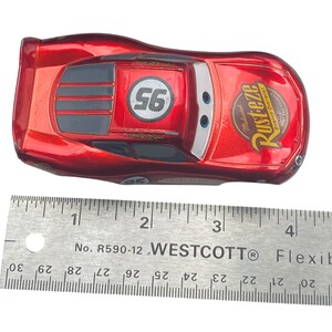 Peut inclure: Une voiture jouet Lightning McQueen rouge avec le num&eacute;ro 95 et la marque Rust-eze. La voiture mesure environ 10 cm de long et a une finition m&eacute;tallique. Le num&eacute;ro 56 est sur le toit. Une r&egrave;gle est au premier plan.