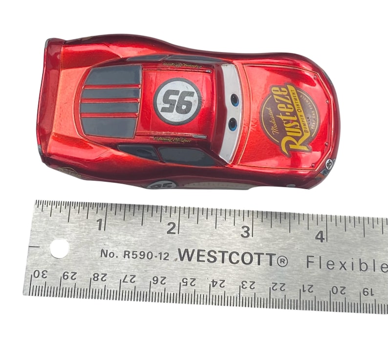 Peut inclure: Une voiture jouet Lightning McQueen rouge avec le num&eacute;ro 95 et la marque Rust-eze. La voiture mesure environ 10 cm de long et a une finition m&eacute;tallique. Le num&eacute;ro 56 est sur le toit. Une r&egrave;gle est au premier plan.