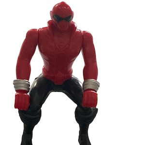 Peut inclure: Une figurine rouge avec un casque et une visière noire, un pantalon noir et des bracelets argentés. La figurine est en position accroupie, les poings serrés. Le jouet est un super-héros ou une figurine d'action.