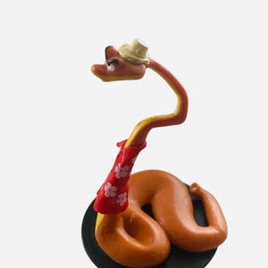 May include: A whimsical figurine of a snake with a yellow and orange body, wearing a small hat and a red scarf adorned with white flower patterns. The snake is coiled on a black base, creating a playful and decorative item.