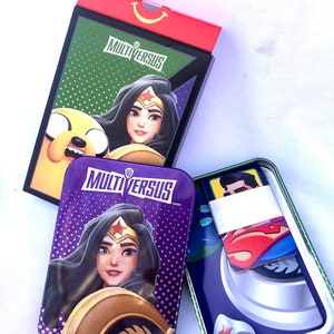 May include: A purple and green tin with a Wonder Woman character on the front. The tin is labeled "MultiVersus" and has a Warner Bros. logo. The tin is open and shows a Superman character card inside.