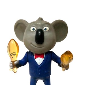 Puede incluir: Un koala gris que lleva un traje azul y una pajarita roja, sosteniendo dos figuritas amarillas en cada mano.