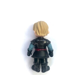 May include: A small plastic figurine of a character from the movie Frozen. The character is wearing a black and blue outfit with a red belt. The figurine is standing on a white background. The text on the back of the figurine reads "Disney Hasbro Frozen 2015."