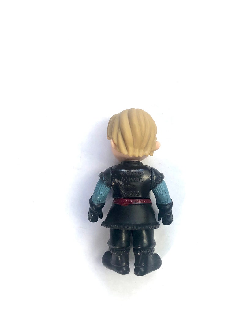 May include: A small plastic figurine of a character from the movie Frozen. The character is wearing a black and blue outfit with a red belt. The figurine is standing on a white background. The text on the back of the figurine reads "Disney Hasbro Frozen 2015."