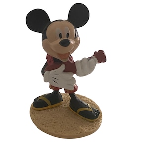 May include: A Mickey Mouse figurine in a dynamic pose. Mickey is wearing red shorts, a red and black jacket, and yellow-trimmed black shoes. His white gloves are outstretched. The figure stands on a textured, tan-coloured base.