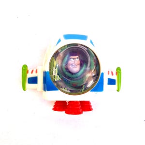 Op de afbeelding: Een Buzz Lightyear speelgoed ruimteschip. De witte behuizing heeft blauwe en groene details, met rode raketboosters. Een doorzichtige koepel toont Buzz' gezicht. De vleugels zijn groen en blauw.