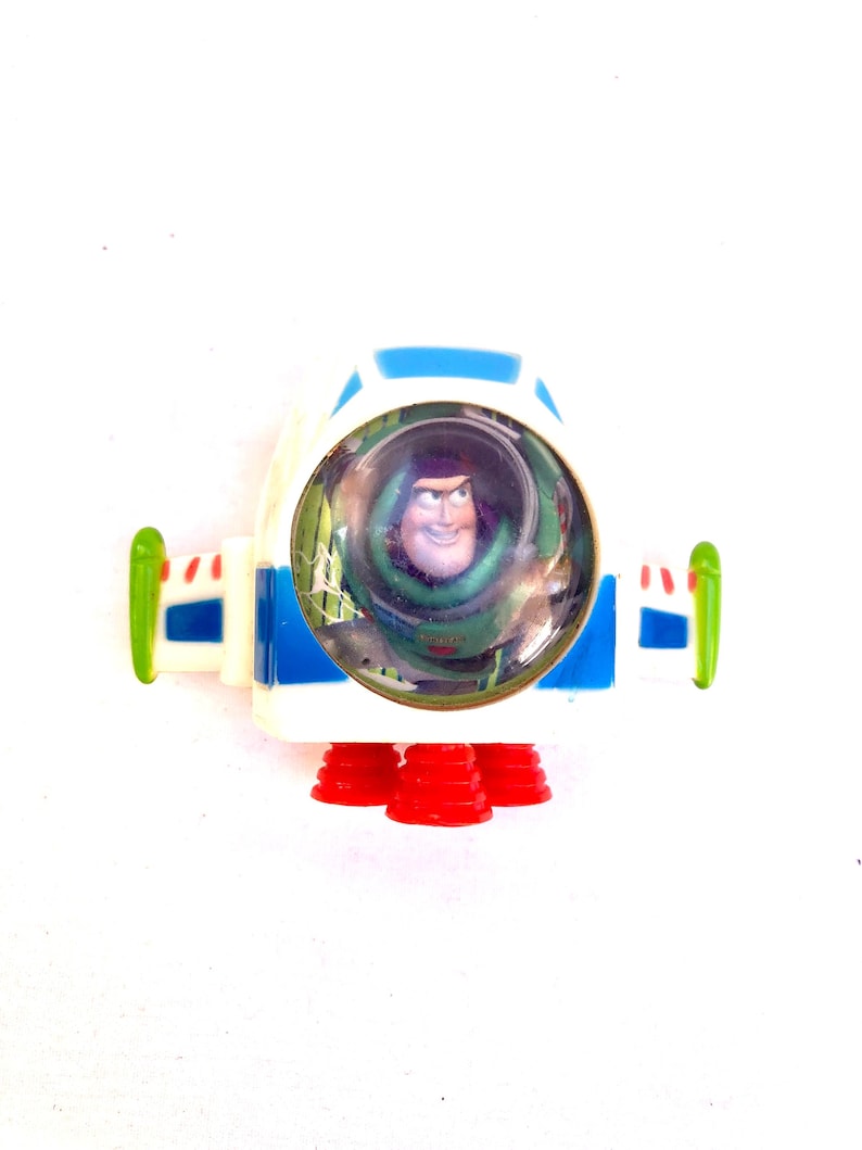 Op de afbeelding: Een Buzz Lightyear speelgoed ruimteschip. De witte behuizing heeft blauwe en groene details, met rode raketboosters. Een doorzichtige koepel toont Buzz' gezicht. De vleugels zijn groen en blauw.
