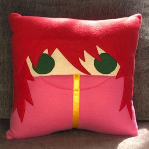 May include: A red and pink plush pillow with a cartoon face. The face has green eyes and a yellow stripe down the centre.
