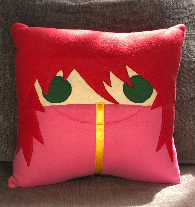 May include: A red and pink plush pillow with a cartoon face. The face has green eyes and a yellow stripe down the centre.