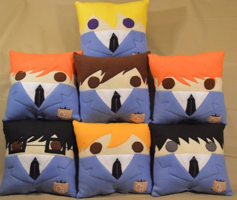 May include: Eight plush pillows featuring characters from the anime Ouran High School Host Club. Each pillow is blue with a white collar and tie, and features a different character's hair and facial features. The pillows are arranged in two rows of four.