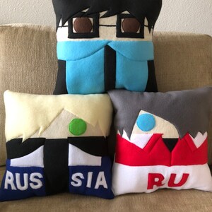 May include: Four plush pillows with cartoon faces and text. The pillows are black, white, grey, and yellow with red, blue, and green accents. The text on the pillows reads "RUSSIA" and "RU".