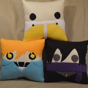May include: Three plush pillows with cartoon faces. The first pillow is white with black eyes and yellow trousers. The second pillow is orange with yellow eyes and a blue shirt. The third pillow is black with grey eyes and a purple scarf.