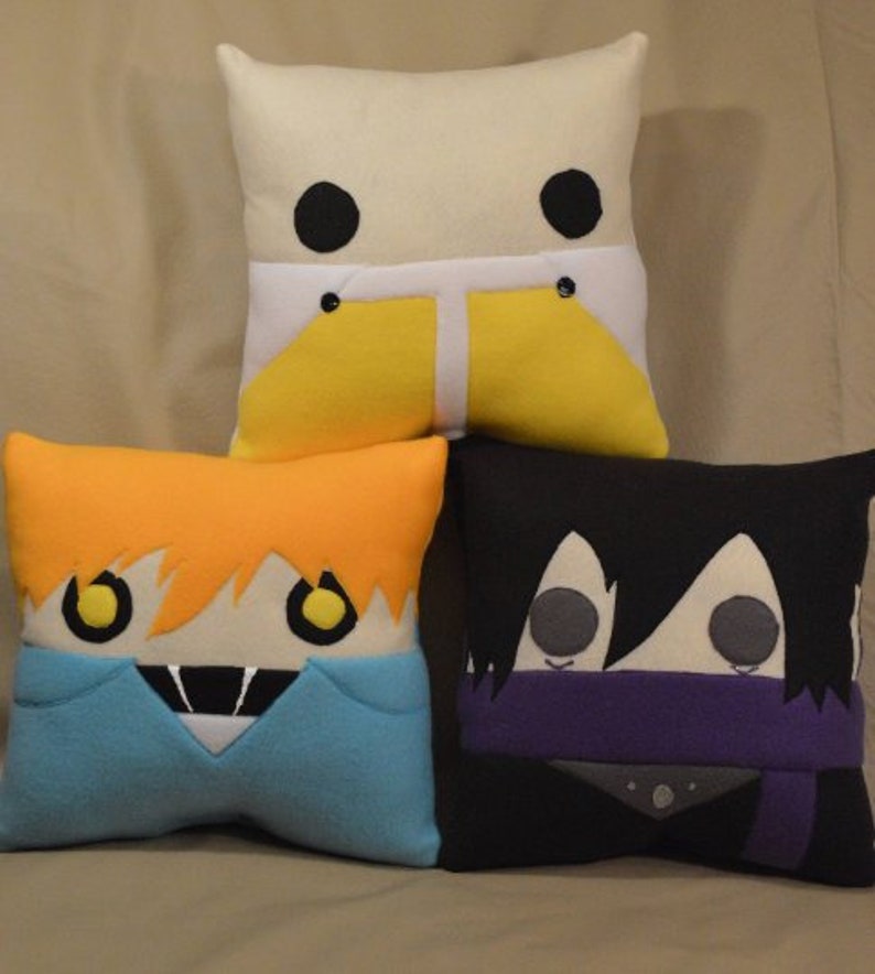 May include: Three plush pillows with cartoon faces. The first pillow is white with black eyes and yellow trousers. The second pillow is orange with yellow eyes and a blue shirt. The third pillow is black with grey eyes and a purple scarf.