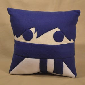 May include: A blue and white plush pillow with a cartoon face. The face has two large eyes and a white mouth.