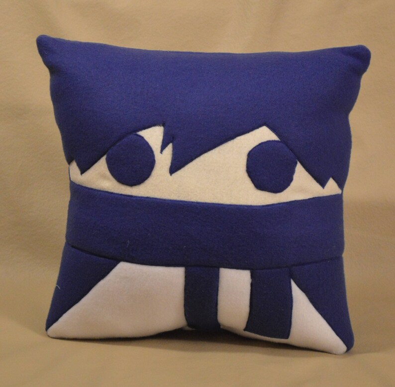 May include: A blue and white plush pillow with a cartoon face. The face has two large eyes and a white mouth.
