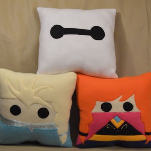 Op de afbeelding: Drie decoratieve kussens met personages uit populaire animatiefilms. Het bovenste kussen is wit met een zwarte omtrek van Baymax uit Big Hero 6. Het linker kussen is crèmekleurig met een blauwe en witte omtrek van Elsa uit Frozen. Het rechter kussen is oranje met een roze, gele en paarse omtrek van Merida uit Brave.