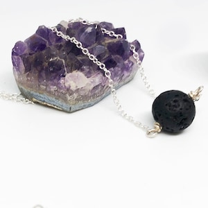 Puede incluir: Un collar de cadena de plata con una cuenta de roca de lava negra. El collar se muestra sobre un lecho de cristales de amatista.