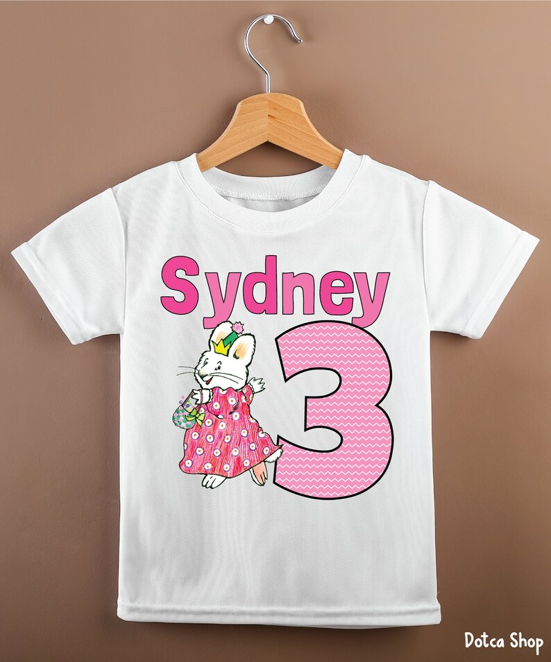 May include: White t-shirt with a pink bunny rabbit wearing a crown and a pink dress with white polka dots. The shirt also has the name "Sydney" and the number "3" in pink.