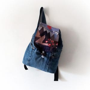 Peut inclure: Un sac à dos en jean bleu avec un rabat en patchwork présentant un motif géométrique dans des tons de brun, violet et rouge. Le rabat a un cercle rouge et une fermeture à cordon blanc.