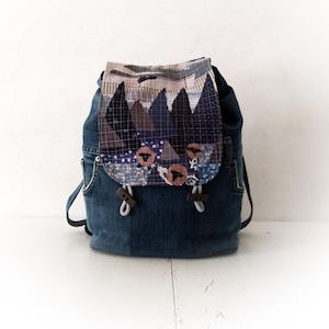 May include: A blue denim backpack with a patchwork flap featuring a geometric pattern in shades of blue, pink, and brown. The flap has a drawstring closure and brown leather accents.