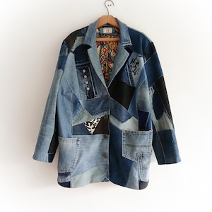 Puede incluir: Blazer vaquero patchwork en varios tonos de azul, con forro estampado de cachemira. La chaqueta presenta múltiples parches de mezclilla, bolsillos y botones. El blazer está colgado en una percha de madera sobre un fondo blanco.