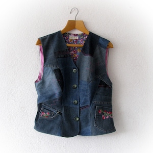 Könnte beinhalten: Eine Patchwork-Jeansweste mit floralem Innenfutter und rosa Nähten. Die Weste hat eine Knopfleiste, Taschen mit floraler Stickerei und hängt an einem Holzbügel vor weißem Hintergrund.
