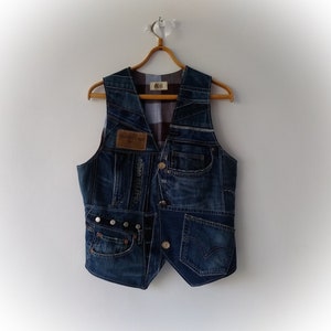May include: A patchwork denim vest made from recycled jeans, in various shades of blue. The vest has pockets, buttons, and a brown leather patch with the text "GRAFTED GOODS". It hangs on a wooden hanger, against a white wall.