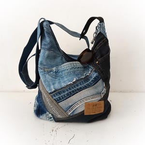 May include: A hobo bag constructed from repurposed blue denim fabric in various shades. The bag features multiple pockets, a dark denim side panel, a brown leather-like patch, and a strap. The text VILA CLOTHES is visible on a grey fabric strip.