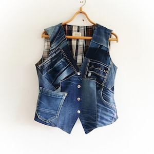 May include: A patchwork denim vest with a V-neck design, featuring a mix of blue jean fabrics. The vest has a plaid lining and is adorned with silver buttons. It hangs on a wooden hanger against a white background.