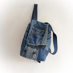 May include: A blue denim backpack with a unique design, crafted from recycled jeans. The bag features multiple pockets, a flap closure, and adjustable straps. The backpack is hanging against a white wall.