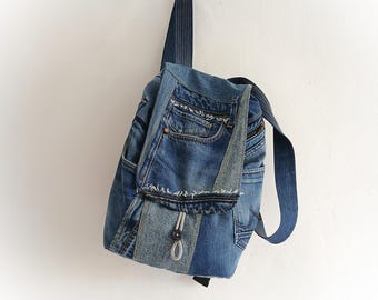 May include: A blue denim backpack with a unique design, crafted from recycled jeans. The bag features multiple pockets, a flap closure, and adjustable straps. The backpack is hanging against a white wall.