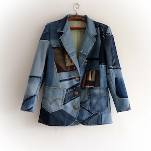 Może przedstawiać: Jeansowa marynarka typu patchwork w różnych odcieniach niebieskiego, wisząca na drewnianym wieszaku. Kurtka posiada liczne naszywki z dżinsu, dwie przednie kieszenie i drewniane guziki. Marynarka jest unikatowa, ręcznie robiona.