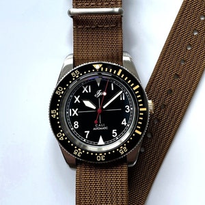 May include: A black and silver wristwatch with a brown nylon strap. The watch face has a black background with white numerals and a gold bezel. The watch face also has the text "CALI AUTOMATIC" printed on it.
