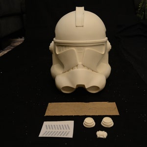May include: A white, unpainted Clone Trooper helmet, a popular item in the Star Wars universe. The helmet is the primary focus, with additional components and a stencil laid out below. The helmet has a distinctive visor and ventilation features.