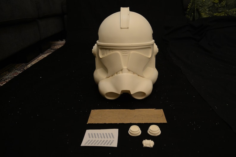 May include: A white, unpainted Clone Trooper helmet, a popular item in the Star Wars universe. The helmet is the primary focus, with additional components and a stencil laid out below. The helmet has a distinctive visor and ventilation features.