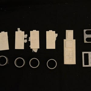 May include: An assortment of off-white plastic model parts, including rectangular pieces with recessed details, cylindrical rings, and two rectangular frames with cut-out shapes. The parts are arranged on a black surface.