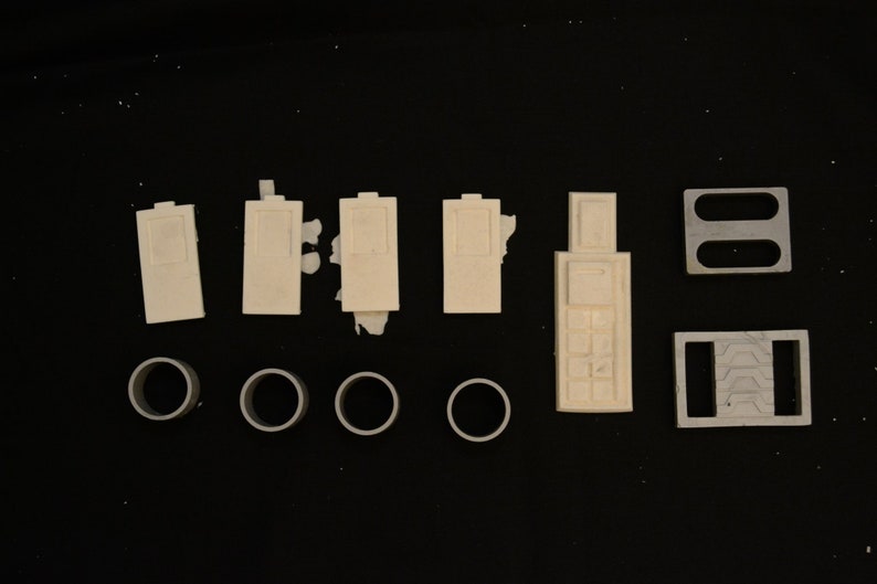 May include: An assortment of off-white plastic model parts, including rectangular pieces with recessed details, cylindrical rings, and two rectangular frames with cut-out shapes. The parts are arranged on a black surface.