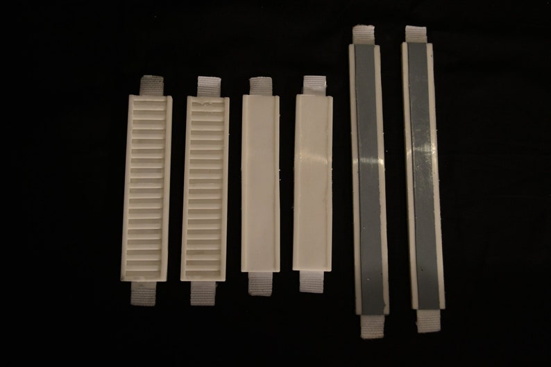 May include: A collection of six white plastic blind slats with fabric straps. Some slats have a slatted design, while others are solid. Two slats have a gray adhesive strip running down the center.
