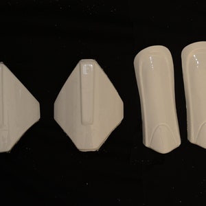 May include: Four pieces of off-white protective gear, including two shin guards and two other pieces, possibly for the chest or shoulders. The items are made of a hard, glossy material and are arranged against a black background.