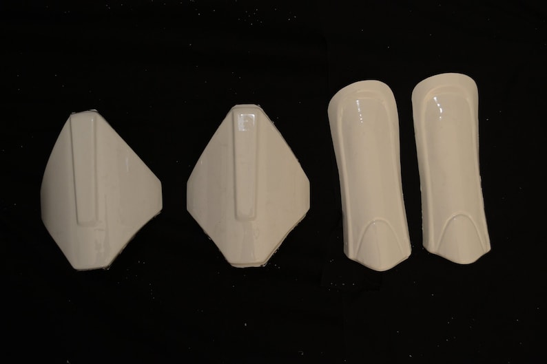 May include: Four pieces of off-white protective gear, including two shin guards and two other pieces, possibly for the chest or shoulders. The items are made of a hard, glossy material and are arranged against a black background.