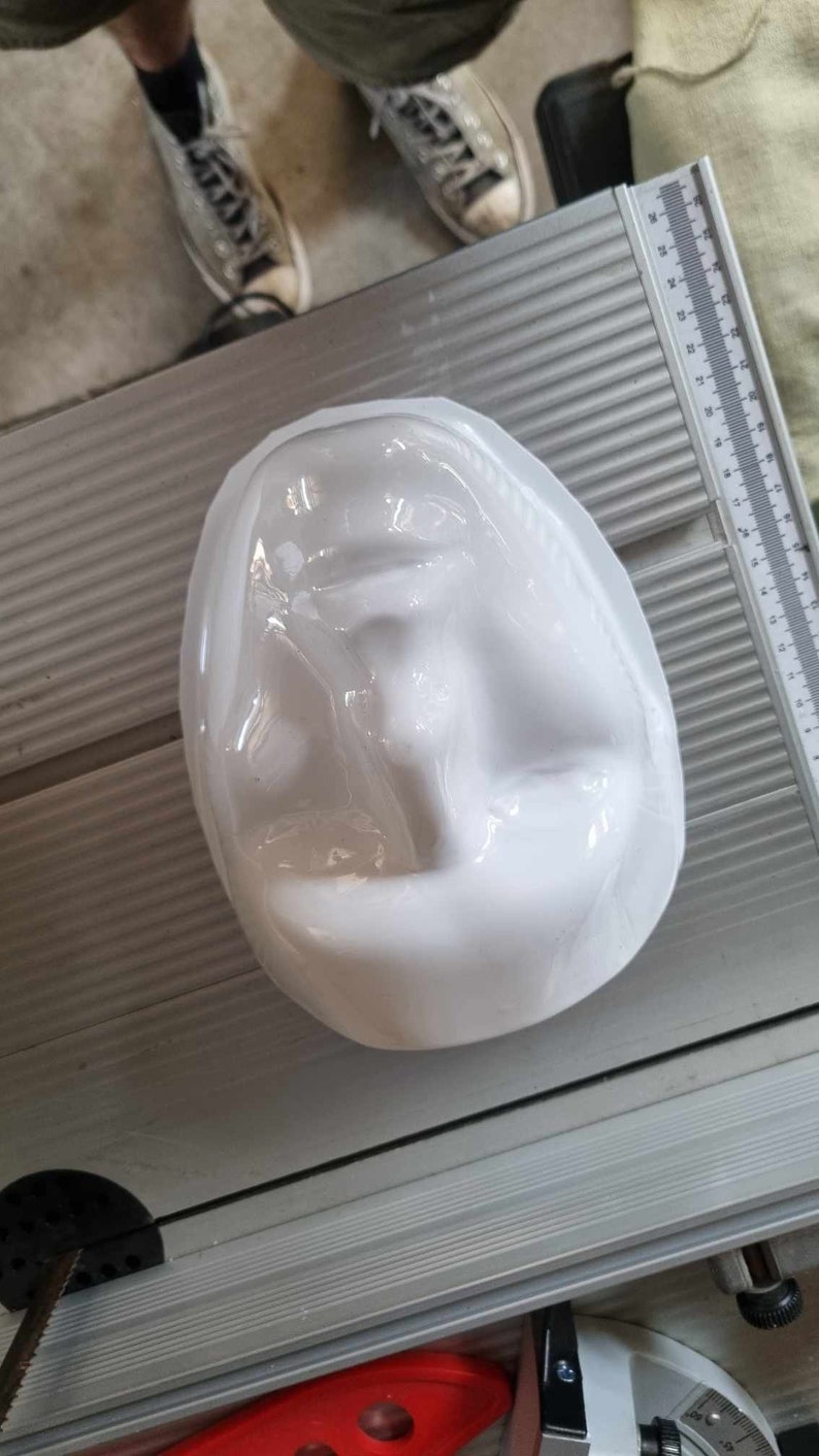 May include: A white, oval-shaped object with a face-like impression. The object is smooth and glossy, possibly made of ceramic or plastic. It sits on a silver surface with a ruler.
