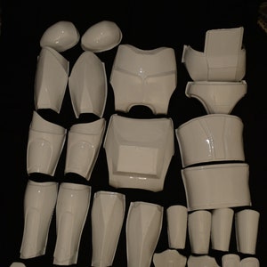 May include: A collection of white, glossy plastic armor pieces, including helmet components, chest plates, shoulder pads, and leg guards. The pieces are arranged on a black surface, suggesting a cosplay or costume set.