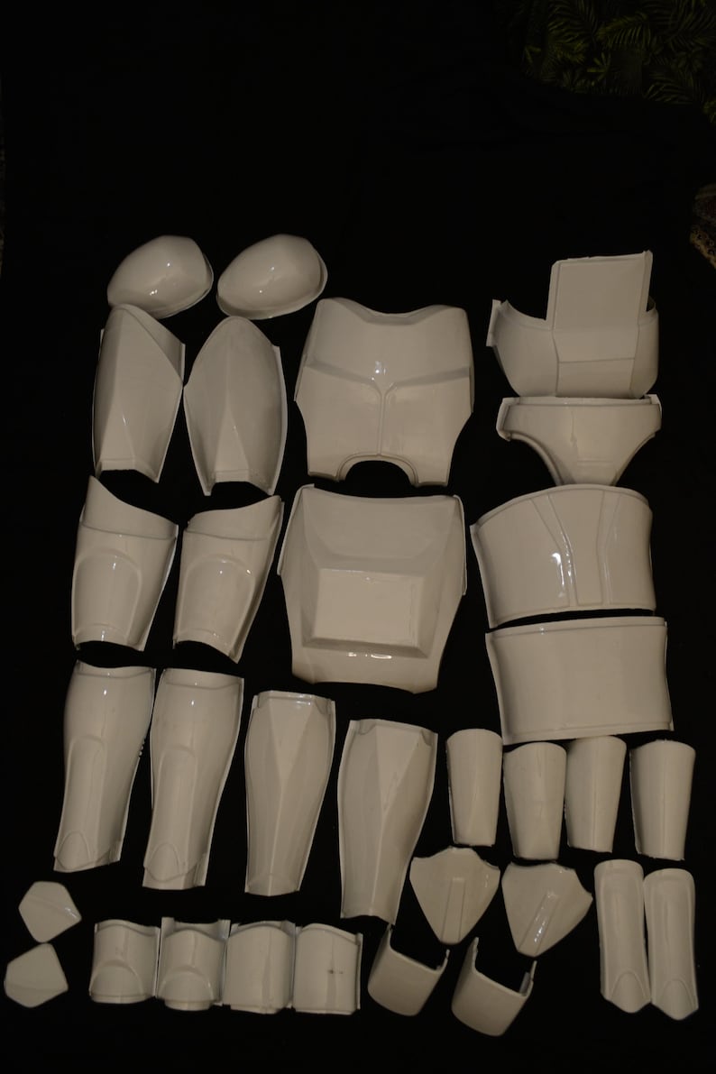 May include: A collection of white, glossy plastic armor pieces, including helmet components, chest plates, shoulder pads, and leg guards. The pieces are arranged on a black surface, suggesting a cosplay or costume set.
