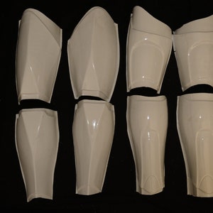 May include: Eight white plastic armor pieces, including shin and thigh guards, are arranged on a black surface. The pieces have a glossy finish and are designed with angular shapes and curves.