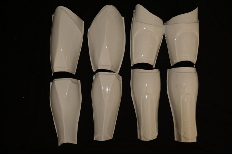 May include: Eight white plastic armor pieces, including shin and thigh guards, are arranged on a black surface. The pieces have a glossy finish and are designed with angular shapes and curves.