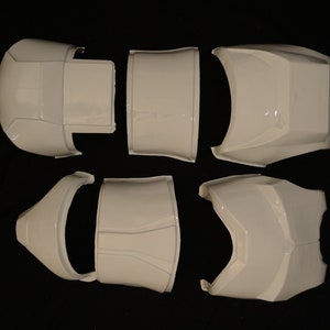 May include: White, segmented armor pieces arranged on a dark surface. The set includes various curved and angular components, likely for a costume or prop. The pieces have a glossy finish and appear to be made of a hard material.